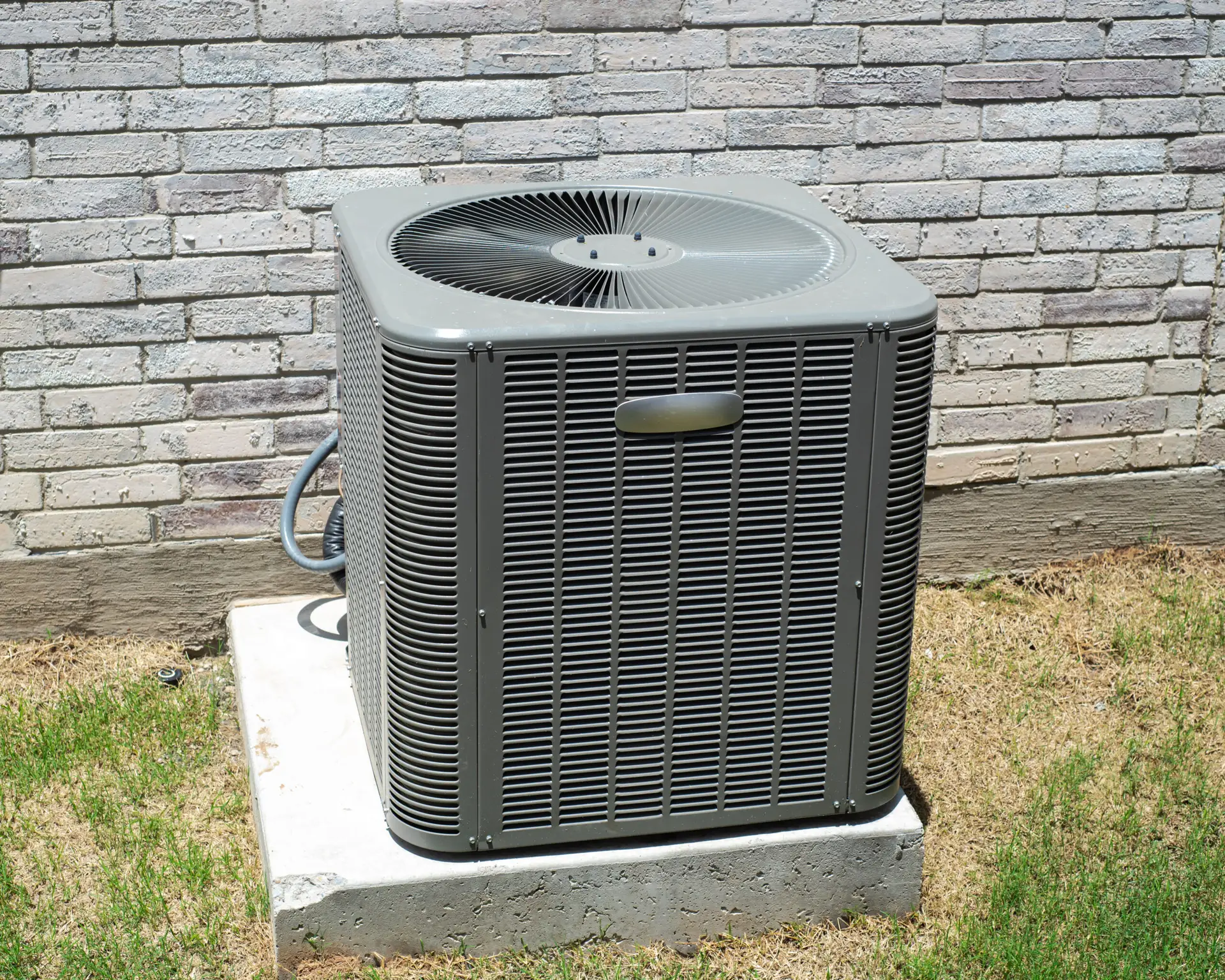 Electrical and refrigerant lines of brand new outdoor central air conditioning unit installed on concrete pad brick house wall. Residential HVAC system, deliver cooled air back into home thru duct. US
